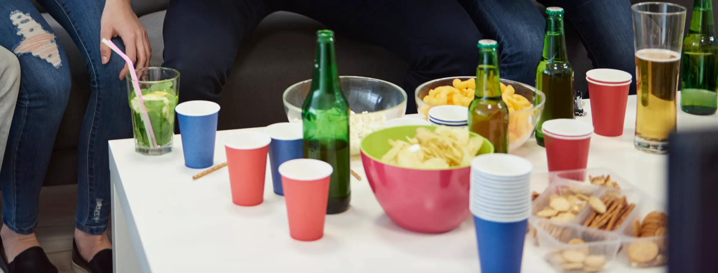 A coffee table set for a game-day party: bowls of chips, cheese puffs and popcorn, trays of pretzels and crackers, bottles, and stacks of red and blue paper cups.