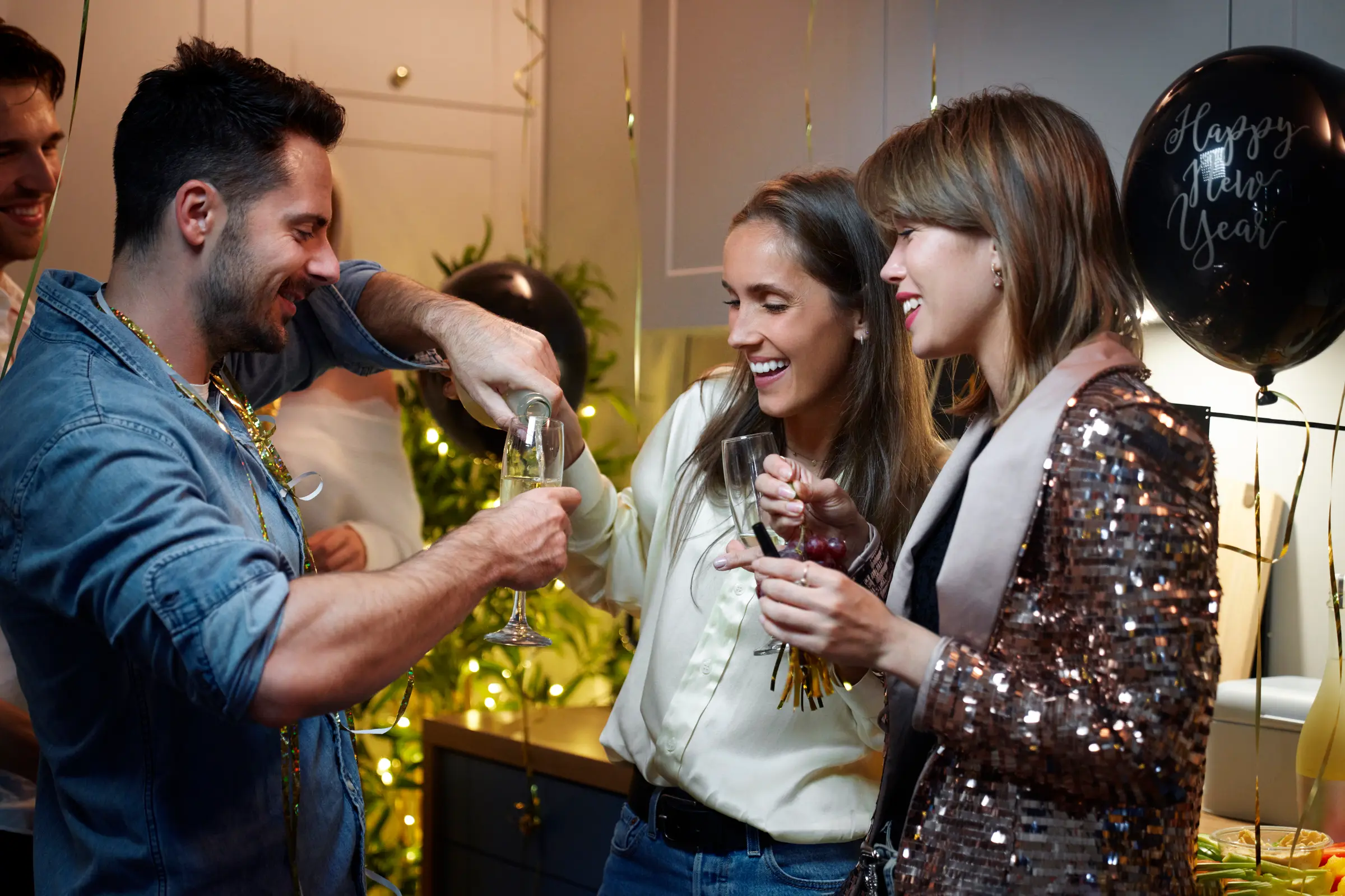 Friends at a house party pouring champagne in a kitchen hung with streamers and balloons