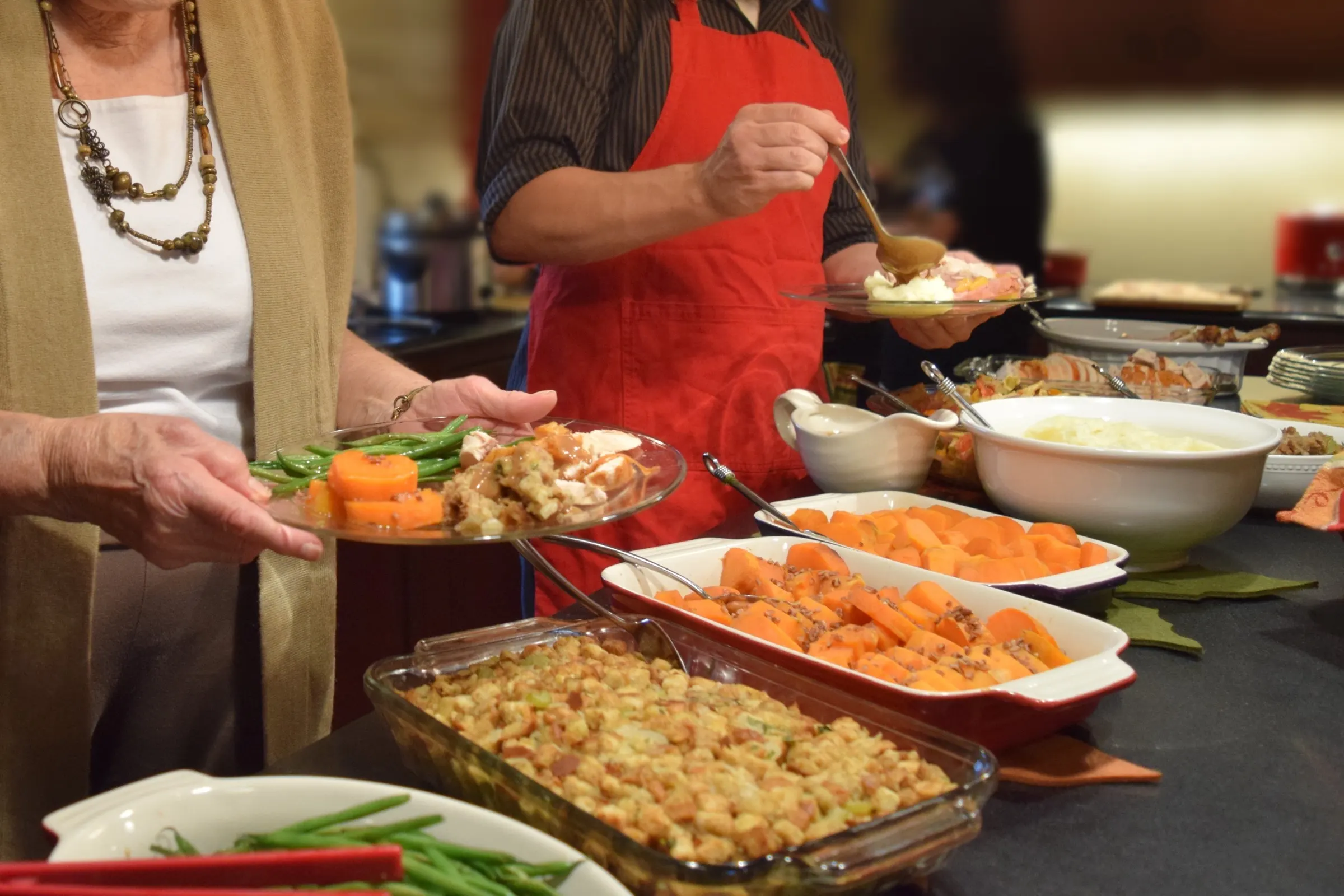 A holiday potluck buffet. One guest holds a plate of green beans, sweet potatoes and stuffing while another, in a red apron, ladles gravy, beside dishes of stuffing, sweet potatoes and mashed potatoes on the counter.