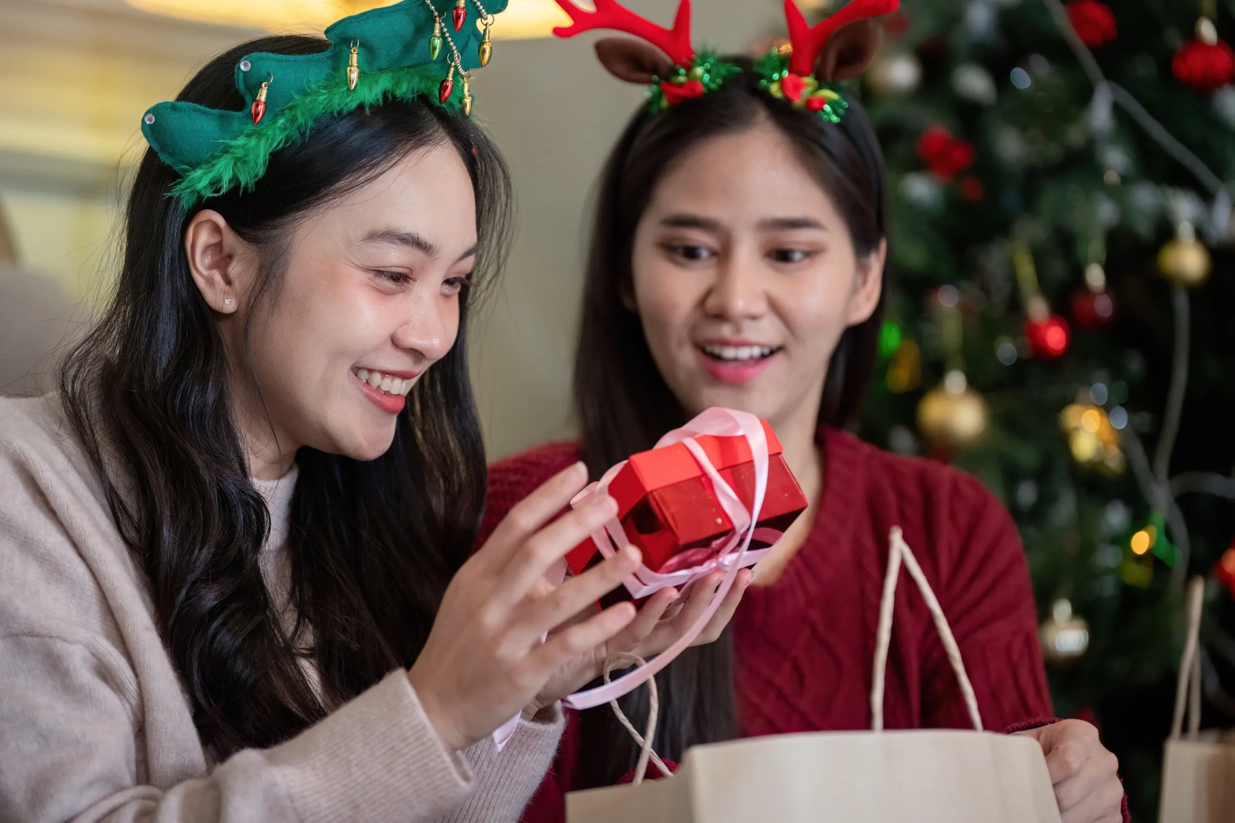 Two friends in holiday headbands grinning as one of them unwraps a small red gift box, a Christmas tree behind them.